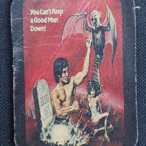 May include: A vintage leather patch with a red background featuring a man fighting a demon with wings. The text "You Can't Keep a Good Man Down!" is at the top of the patch. The man is standing on a tombstone that reads "Bruce Lee".