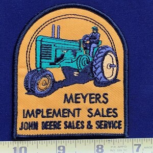 May include: A yellow and green embroidered patch with a cartoon image of a John Deere tractor. The text on the patch reads "Meyers Implement Sales John Deere Sales & Service".