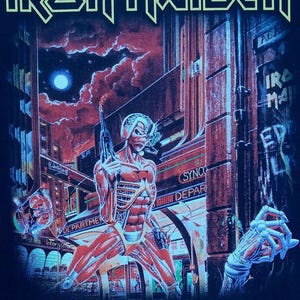 Könnte beinhalten: Schwarzes T-Shirt mit dem Bandnamen "Iron Maiden" in gelben Buchstaben. Die Grafik zeigt eine Cyborg-Figur mit einer Waffe vor einer futuristischen Stadtkulisse. Der Text "SOMEWHERE IN TIME" steht darunter.
