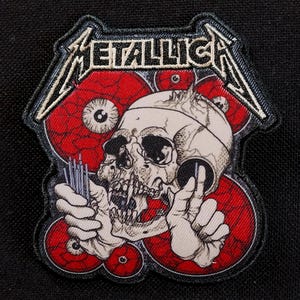 May include: Embroidered patch with the word "METALLICA" in white, above a skull and red eye designs. The patch has a black border, suitable for application on clothing or accessories.