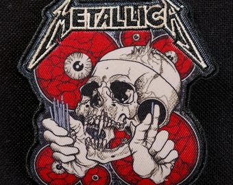 May include: Embroidered patch featuring the band name "METALLICA" in white, above a skull with red eye designs. The patch has a black border and is likely for clothing or accessories.