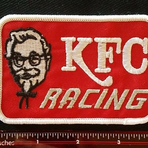 Op de afbeelding: Een geborduurde patch in rood en wit met de tekst "KFC Racing" en een cartoonafbeelding van Colonel Sanders.