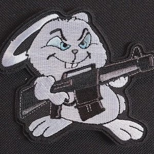 May include: Embroidered patch featuring a cartoon rabbit with a mischievous expression, holding a machine gun. The rabbit is primarily gray and white, with blue eyes and a black outline. The patch has a black border and is set against a black background.