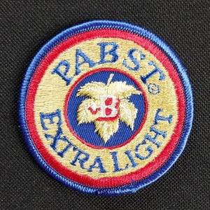 May include: A round, blue, red and gold embroidered patch with the words "Pabst Extra Light" and a stylised hop leaf with a red "B" in the centre.