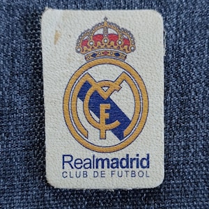May include: A white patch with a gold and blue Real Madrid crest, a crown above the crest, and the text "Realmadrid CLUB DE FUTBOL" below the crest.