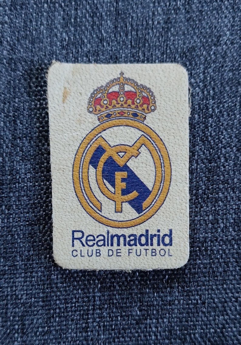 May include: A white patch with a gold and blue Real Madrid crest, a crown above the crest, and the text "Realmadrid CLUB DE FUTBOL" below the crest.