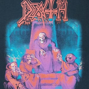Puede incluir: Camiseta negra con un diseño gráfico de la banda Death. El diseño incluye el logotipo naranja de la banda y una escena con figuras con túnicas y un esqueleto. El texto "SCREAM BLOODY GORE" está en la parte inferior.