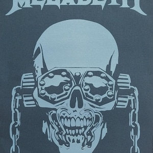Könnte beinhalten: Graue Grafik mit dem Wort "MEGADETH" über einem Schädel mit Brille und Ketten. Der Schädel hat ein detailliertes Design mit sichtbaren Zähnen und einem metallischen Aussehen. Der Hintergrund ist dunkelgrau.