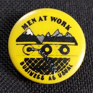 Può includere: Bottone giallo con testo nero che recita "Men at Work Business as Usual". Il bottone presenta un'illustrazione in bianco e nero di una catena montuosa, una catena e due ruote.