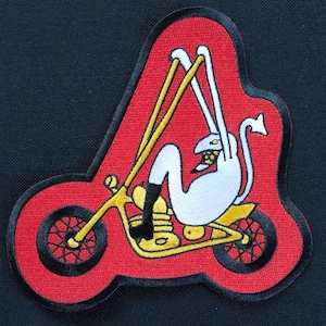 Op de afbeelding: Een geborduurde patch in rood en zwart, met een witte cartoonfiguur die op een gele motor rijdt. Het figuur heeft een lange staart en draagt een zwarte helm.