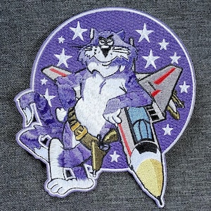 Puede incluir: Parche bordado con un gato de dibujos animados en morado y blanco, sosteniendo un misil. El gato está sobre un fondo morado con estrellas blancas. El parche tiene un diseño de estilo militar.
