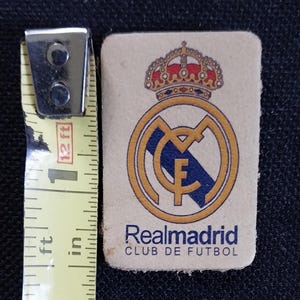 May include: A white leather patch with a gold and blue Real Madrid logo. The logo features a crown, a soccer ball, and the text "Realmadrid CLUB DE FUTBOL".