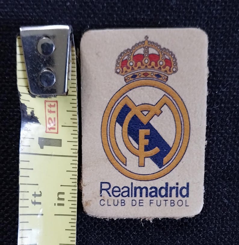 May include: A white leather patch with a gold and blue Real Madrid logo. The logo features a crown, a soccer ball, and the text "Realmadrid CLUB DE FUTBOL".