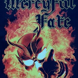 May include: Black t-shirt featuring a fiery skull with horns and the band name "Mercyful Fate" in white gothic lettering. The image includes the text "Don't break the oath" at the bottom, with a hand pointing forward.