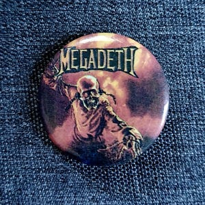 May include: A round button with the word "MEGADETH" in a black, gothic font. The button features a skull-like figure with a raised arm against a dark, reddish-brown background. The button is set against a textured, dark blue fabric.