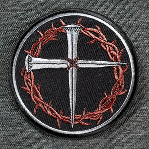 May include: Round black patch with a white border featuring a silver cross made of nails, encircled by a red and brown thorny wreath. The patch is embroidered with detailed stitching.