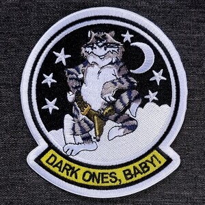 Könnte beinhalten: Gestickter Aufnäher mit einem Cartoon-Katzen-Design, das ein goldenes Objekt hält, vor einem Nachthimmel mit Sternen und einem Halbmond. Der Aufnäher trägt den Text "DARK ONES, BABY!" in Gelb auf schwarzem Hintergrund mit weißem Rand.
