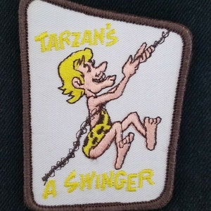 May include: A white embroidered patch with a brown border. The patch features a cartoon image of Tarzan swinging on a vine. The text "Tarzan's A Swinger" is written in yellow lettering.