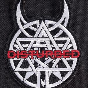 May include: Embroidered patch with a white geometric design and red "DISTURBED" text. The patch has a black border and a horned shape at the top. Suitable for application on clothing or bags.
