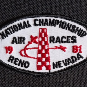 Könnte beinhalten: Weißer Patch mit schwarzem gesticktem Text "National Championship Air Races 1981 Reno Nevada" und einem rot-weiß karierten Turm mit einem roten Pfeil, der nach rechts zeigt.