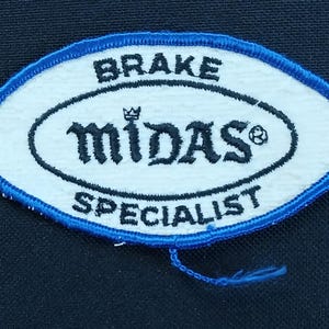 May include: Oval-shaped patch with a white center and a blue border. The patch features the words "BRAKE" and "SPECIALIST" in black, with "midas" in a stylized font. A small crown and a soccer ball icon are also present.