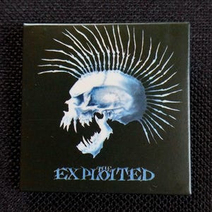 Pode incluir: Botão quadrado preto com um desenho de caveira azul e branco com moicano. A caveira tem a boca aberta com dentes afiados. O texto "EXPLOITED" está escrito em azul na parte inferior do botão.