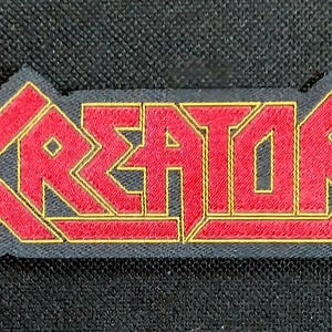 Könnte beinhalten: Gestickter Aufnäher mit dem Wort "KREATOR" in Rot, gold umrandet, auf schwarzem Hintergrund. Der Aufnäher hat eine rechteckige Form mit gezacktem Rand. Der Text ist in einer fetten, stilisierten Schriftart.