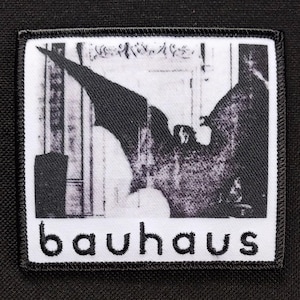 May include: A rectangular patch with a black and white graphic of a figure with outstretched wings, and the word "bauhaus" embroidered in black on a white background. The patch has a black border and is set against a black background.