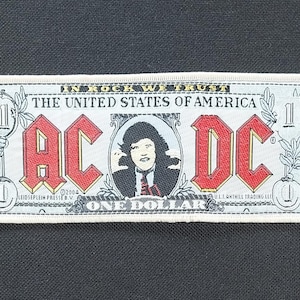 Puede incluir: Un parche rectangular blanco que imita un billete de un dólar, con el logotipo de AC/DC en letras rojas. El texto incluye "IN ROCK WE TRUST" y "THE UNITED STATES OF AMERICA". Un retrato de una persona está en el centro. Fondo negro.