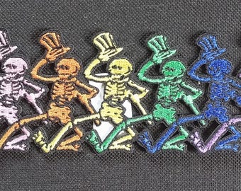 以下が含まれることがあります： ラベンダー、オレンジ、黄色、緑、青、紫の様々な色の、6つの刺繍されたスケルトンパッチの列。 各スケルトンはシルクハットをかぶり、踊っています。 背景は黒です。