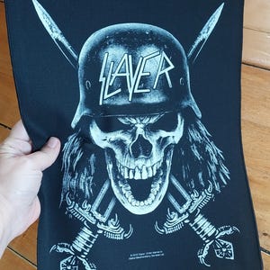 May include: Black patch featuring a skull wearing a helmet with the word "SLAYER" in white. Two crossed swords are behind the skull. The patch is rectangular and made of fabric.