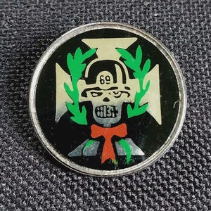 May include: Round metal pin with a black background and a silver border. The pin features a skull with the number "69" above it, a green laurel wreath, and a red bow tie. A silver cross shape is behind the skull.