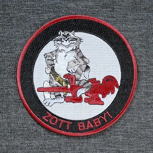 May include: Embroidered patch with a cartoon cat design. The cat is grey and white, holding a golden object, and seated on a red plane. The patch has a black border with a red outline and the text "ZOTT BABY!" in red.