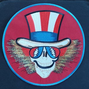 May include: A red, white, and blue embroidered patch of a skull wearing a top hat and sunglasses. The skull is smiling and has a long, white beard. The patch is circular and has a blue border.