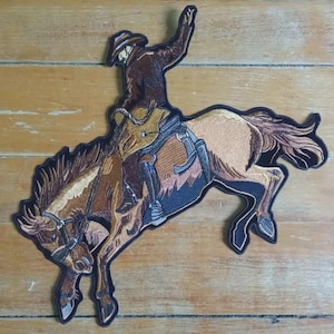Pode incluir: Patch bordado com um cowboy montando um cavalo selvagem. O patch tem um esquema de cores marrom, bege e preto. O cowboy está usando um chapéu e jaqueta, com o braço levantado. O cavalo está empinando. Tema ocidental.