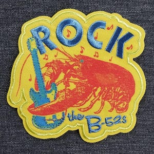 Puede incluir: Parche bordado con fondo amarillo, con una langosta roja tocando una guitarra azul. La palabra "ROCK" es azul, con notas musicales. El texto "the B-52s" es azul.