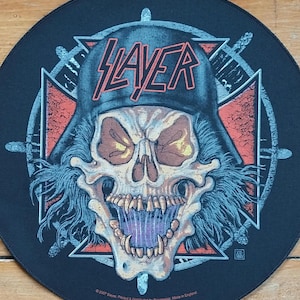May include: Round black patch featuring a skull wearing a helmet with the word "SLAYER" in red letters. The skull has yellow eyes and blue hair. The patch has a red and white cross design.