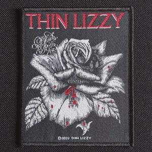 May include: Black rectangular patch featuring the text "THIN LIZZY" in red above a detailed black and white rose with red accents. The patch includes the text "©2022 THIN LIZZY".