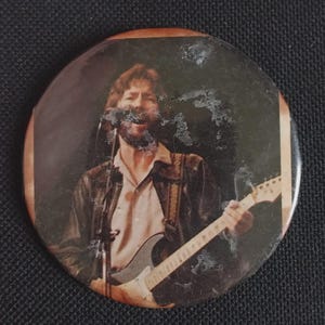 May include: Round button featuring a musician playing a guitar. The button has a brown border and a faded image of a man with a beard, wearing a leather jacket and holding a guitar. The background is dark.