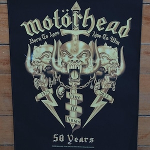 May include: Black rectangular graphic featuring the band Motörhead's logo in gold. The design includes a skull with tusks, a sword, and the text "Born To Lose, Live To Win." It also says "50 Years" and the years 1975 and 2025.