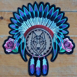 May include: An embroidered iron-on patch featuring a wolf's head inside a dreamcatcher with feathers and roses. The wolf is wearing a headdress with blue, purple, and white feathers.