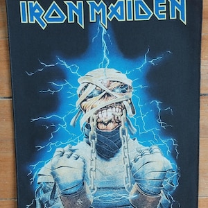 Puede incluir: Parche rectangular negro con el nombre de la banda "IRON MAIDEN" en letras azules delineadas. La imagen muestra una figura momificada con cadenas, rodeada de rayos azules. El nombre de la banda está en la parte superior.