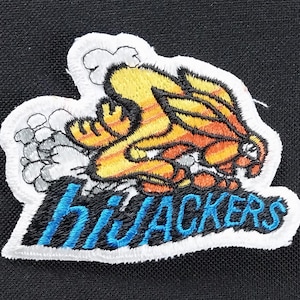 May include: White embroidered patch featuring a cartoon orange and yellow creature with the word "hijackers" embroidered in blue and white underneath.