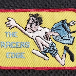 May include: A yellow embroidered patch with red stitching featuring a cartoon character running with a blue denim jacket. The text "THE RACERS EDGE" is embroidered in blue on the patch.