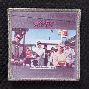 May include: A square patch with a silver border features a band photo with the text "AC/DC" in pink above the image. The image shows a group of people with black bars over their eyes. Below the image is the text "Dirty Deeds Done Dirt Cheap."