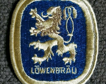 May include: Embroidered patch with a gold lion rampant on a blue field, the word "LÖWENBRÄU" in white text below. The patch has a gold border and a rectangular shape with rounded corners. A vintage style patch.