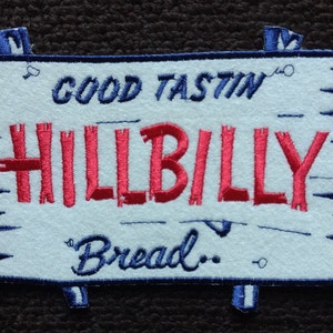 Può includere: Una toppa ricamata bianca con cuciture blu. La toppa ha la forma di un cartello di legno con un bordo sfrangiato. Il testo sulla toppa recita "Good Tastin' Hillbilly Bread."