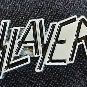 May include: A silver-colored metal pin with the word "SLAYER" in a stylized, bold font. The letters have a black outline and a reflective silver interior. The pin is set against a dark, textured background.