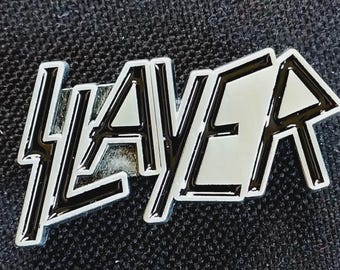 May include: A silver-colored metal pin with the word "SLAYER" in a stylized, bold font. The letters have a black outline and a reflective silver interior. The pin is set against a dark, textured background.