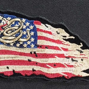 May include: A distressed American flag patch with a snake design in the upper left corner. The flag is frayed and torn around the edges, giving it a vintage look.